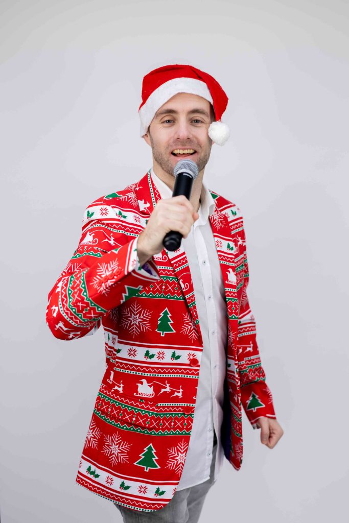 Dennis in een rode kerstoutfit met kerstmuts en microfoon.