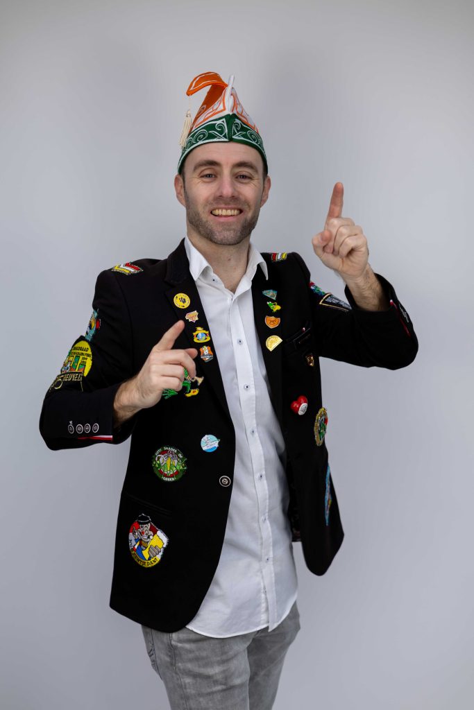 Dennis in carnavalsjas met emblemen en een groene feesthoed.