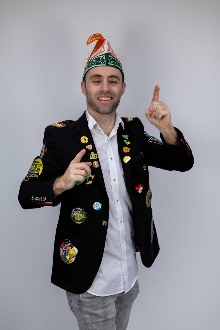 Dennis in carnavalsjas met emblemen en een groene feesthoed.