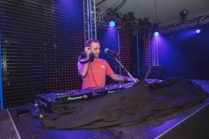 DJ Dennis met microfoon achter de draaitafel, omringd door paarse feestverlichting.