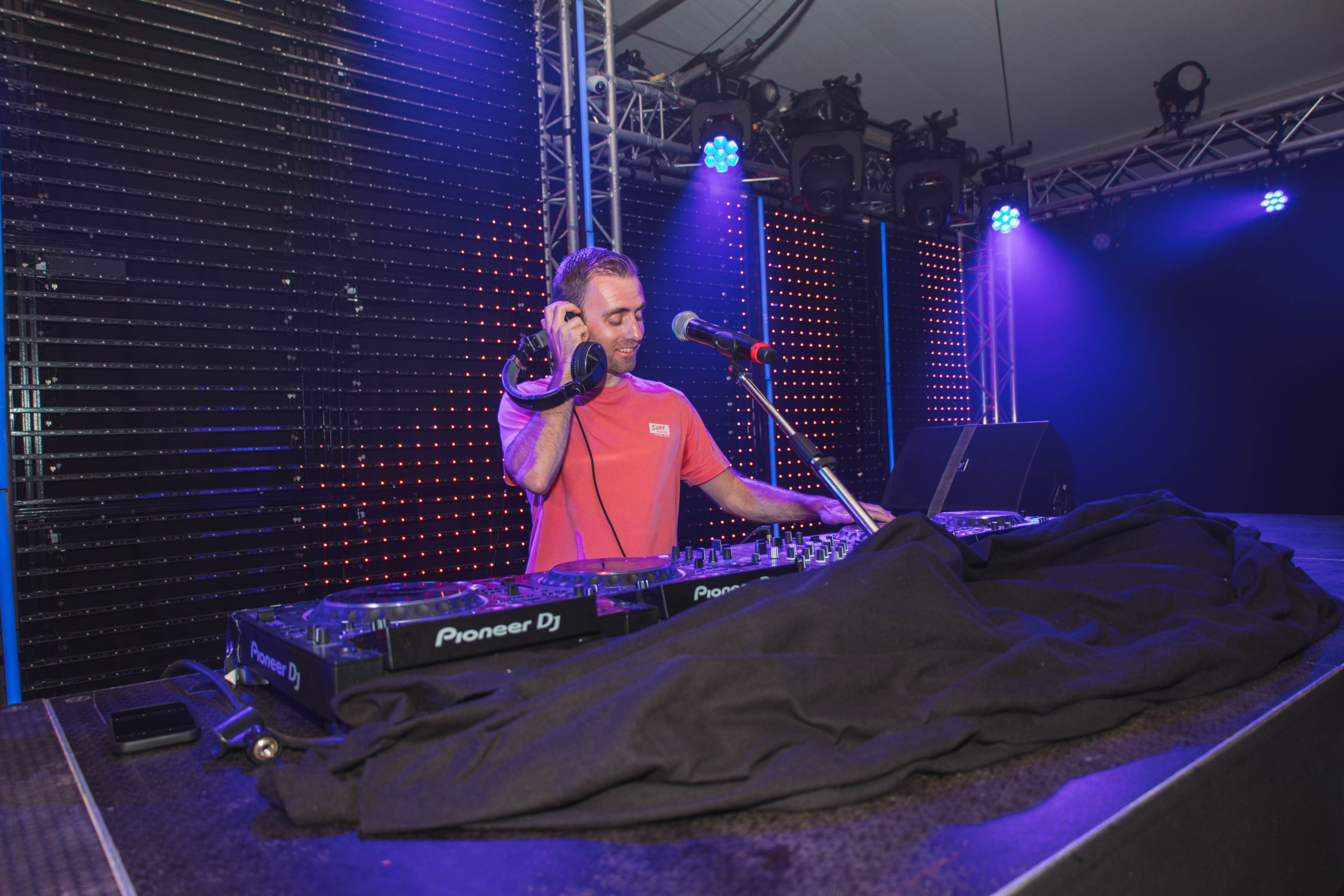 DJ Dennis met microfoon achter de draaitafel, omringd door paarse feestverlichting.