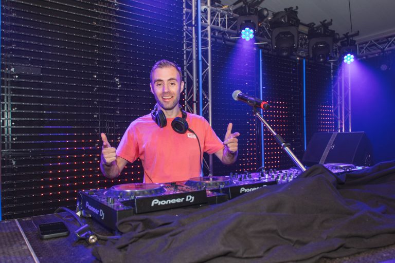 DJ Dennis in een rood shirt met koptelefoon achter de DJ-booth.
