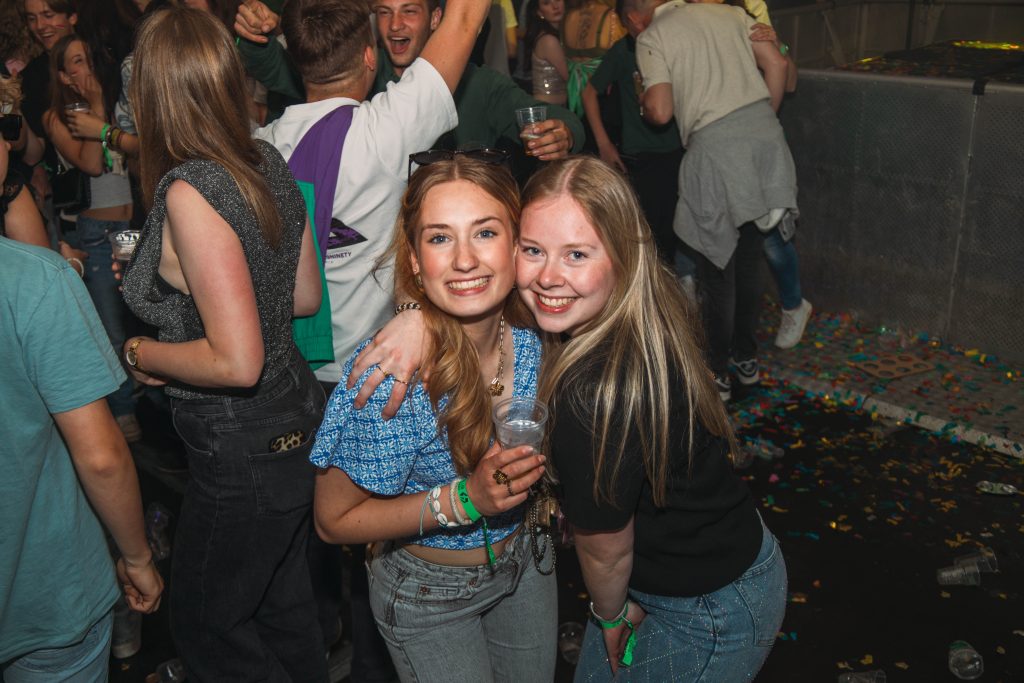Twee feestende gasten lachen naar de camera op een drukke dansvloer.