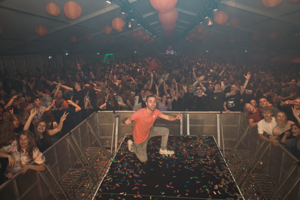 DJ Dennis bij de draaitafel voor een volle zaal met feestende gasten.