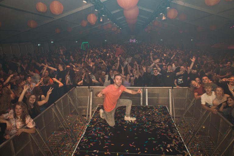 DJ Dennis bij de draaitafel voor een volle zaal met feestende gasten.