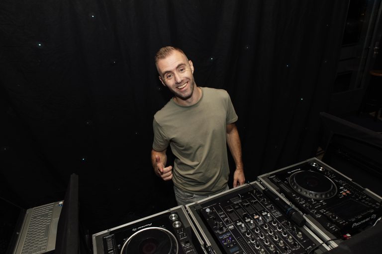 DJ Dennis lacht achter zijn DJ-apparatuur en draagt een groen T-shirt.