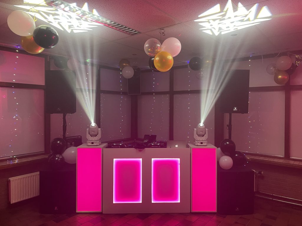 DJ-meubel en speakers met roze verlichting in een zaal met ballonnen.