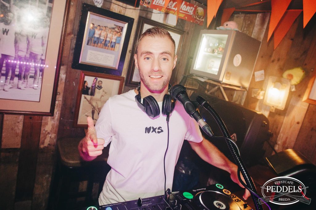 DJ Dennis lacht achter de DJ-apparatuur in een café met sfeerverlichting.