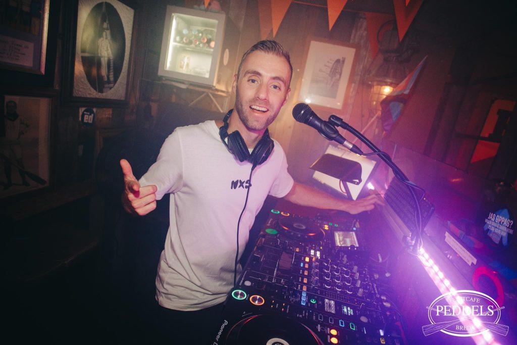 DJ Dennis met microfoon en koptelefoon achter de draaitafel tijdens een optreden.