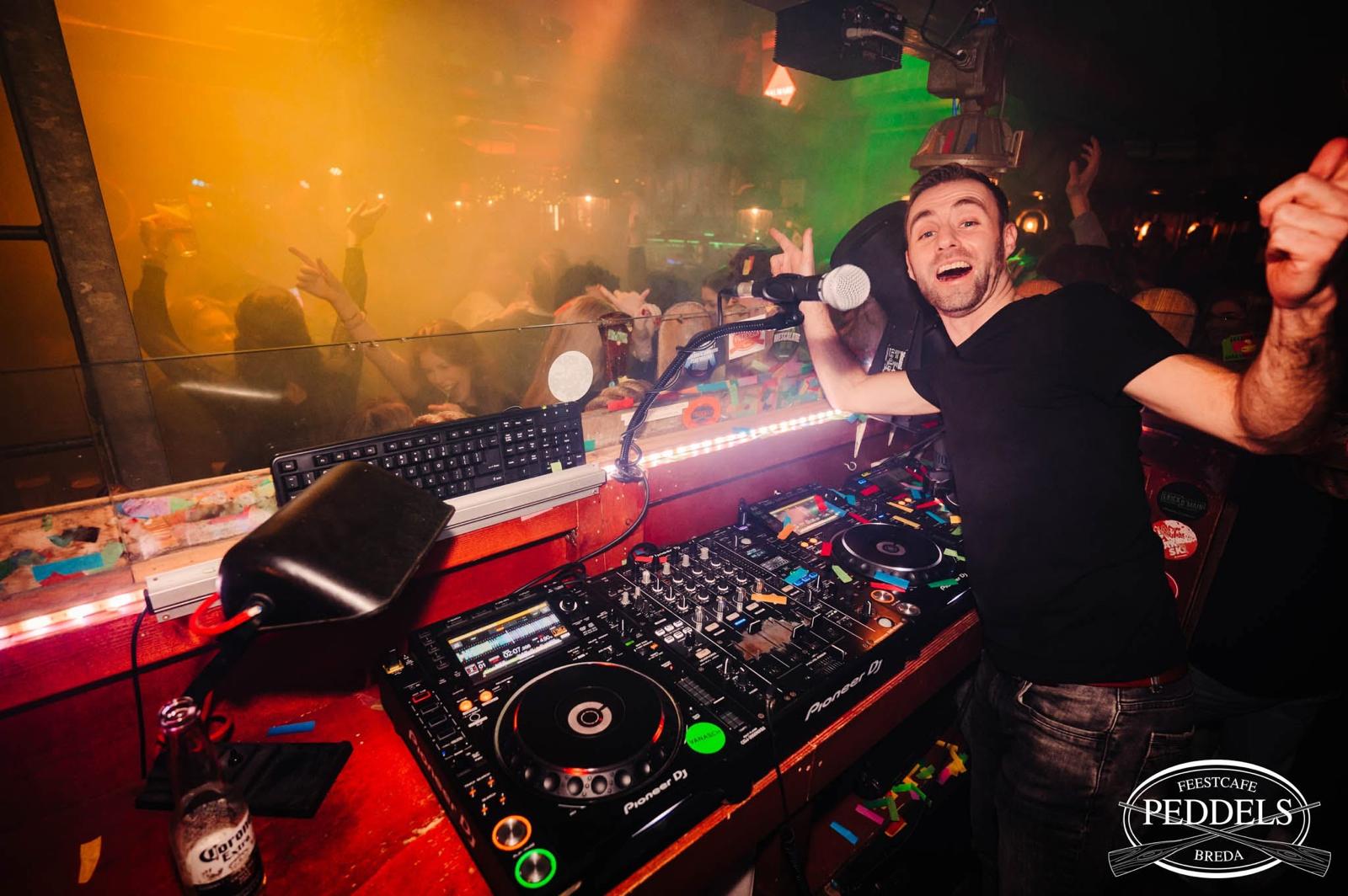 DJ Dennis met zijn arm in de lucht achter de draaitafel, tussen kleurrijk licht en feestend publiek.