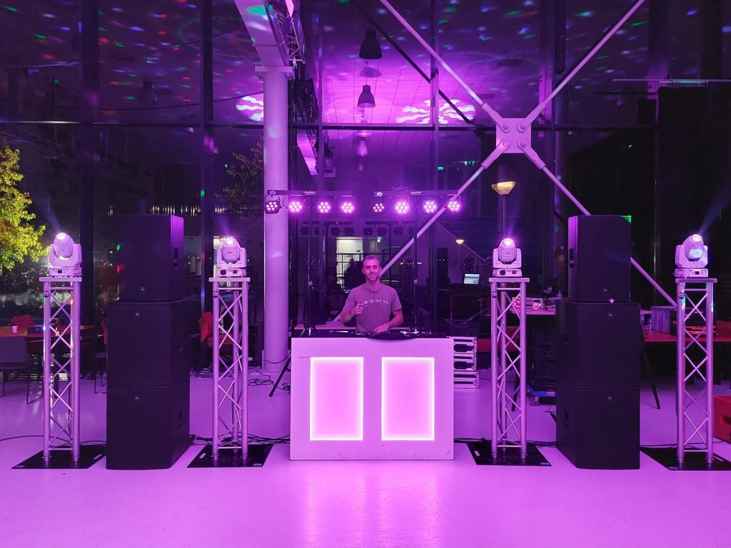 Dennis achter een wit verlicht DJ-meubel met vier trussen, movingheads en grote speakers.