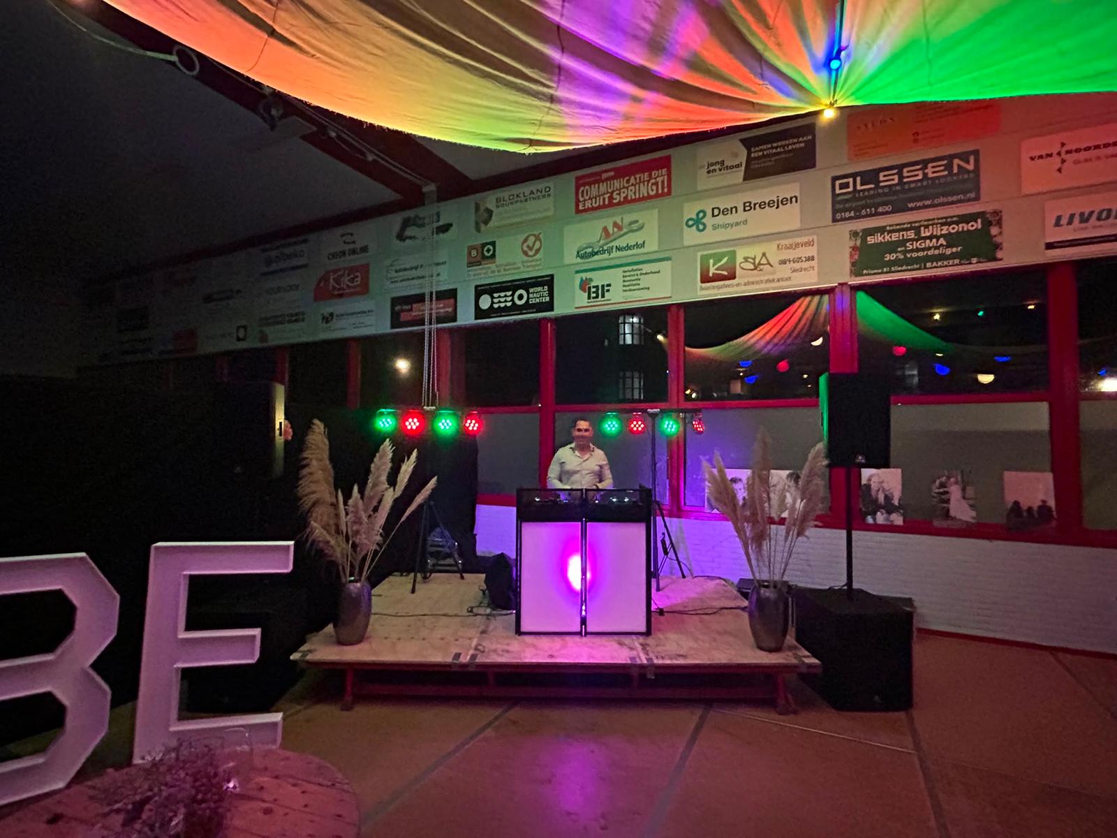 Dennis achter een verlicht DJ-meubel met LED-spots op een feestlocatie.